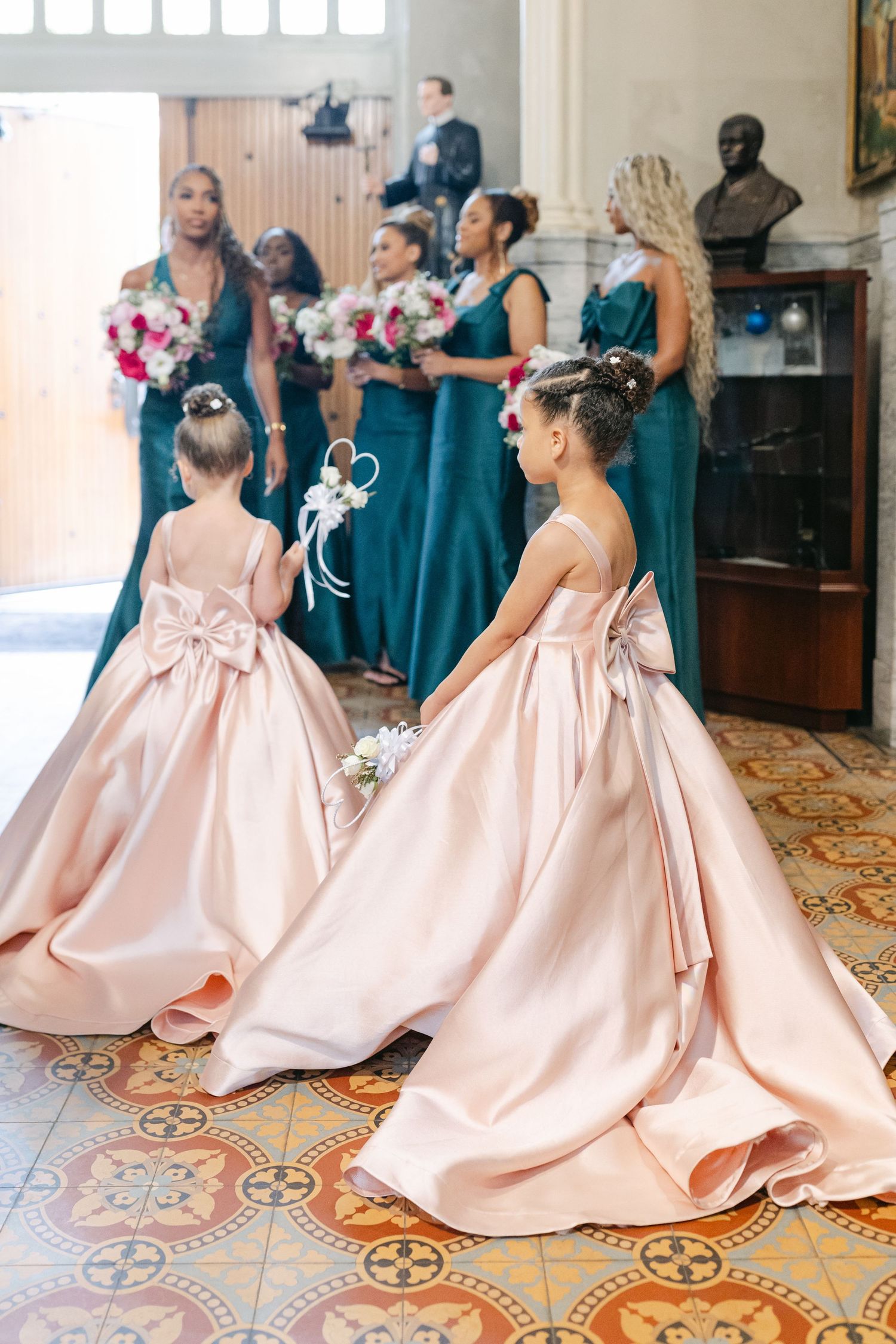 Young bridesmaids in matching pink satin dresses twirl and play on ornate tiled floor at elegant wedding venue.