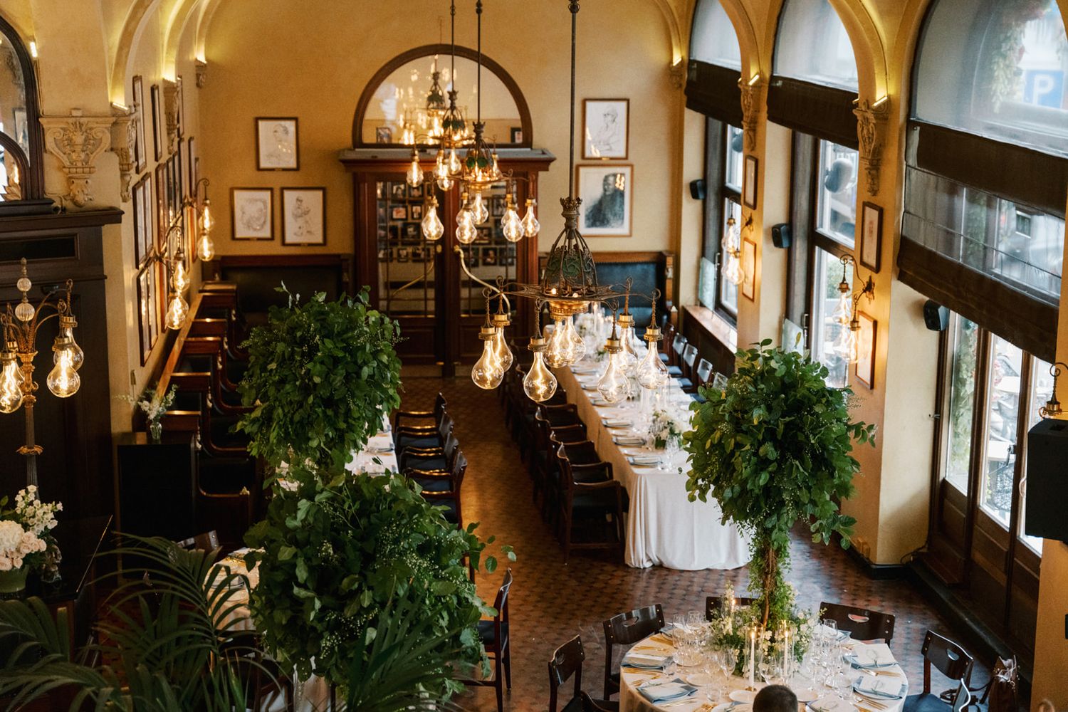 Elegant restaurant interior with chandeliers, arched windows, and lush greenery creating a sophisticated dining atmosphere.