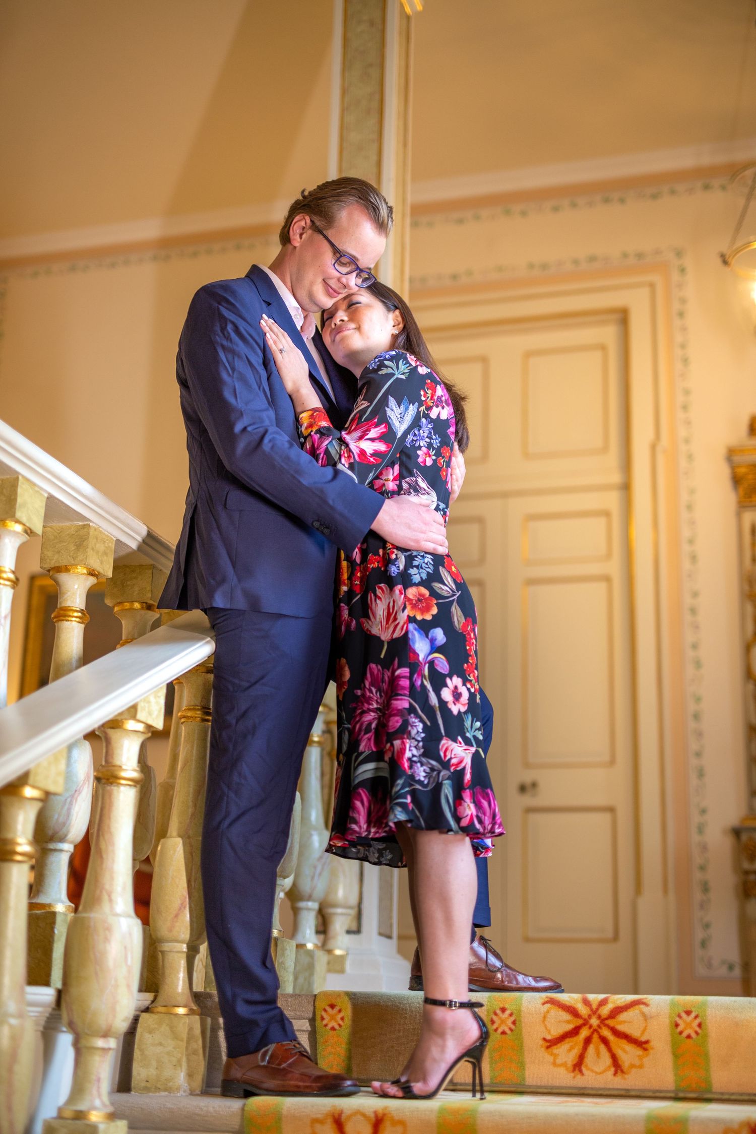 Romantic couple embraces on elegant staircase with golden banister in formal attire during special occasion.