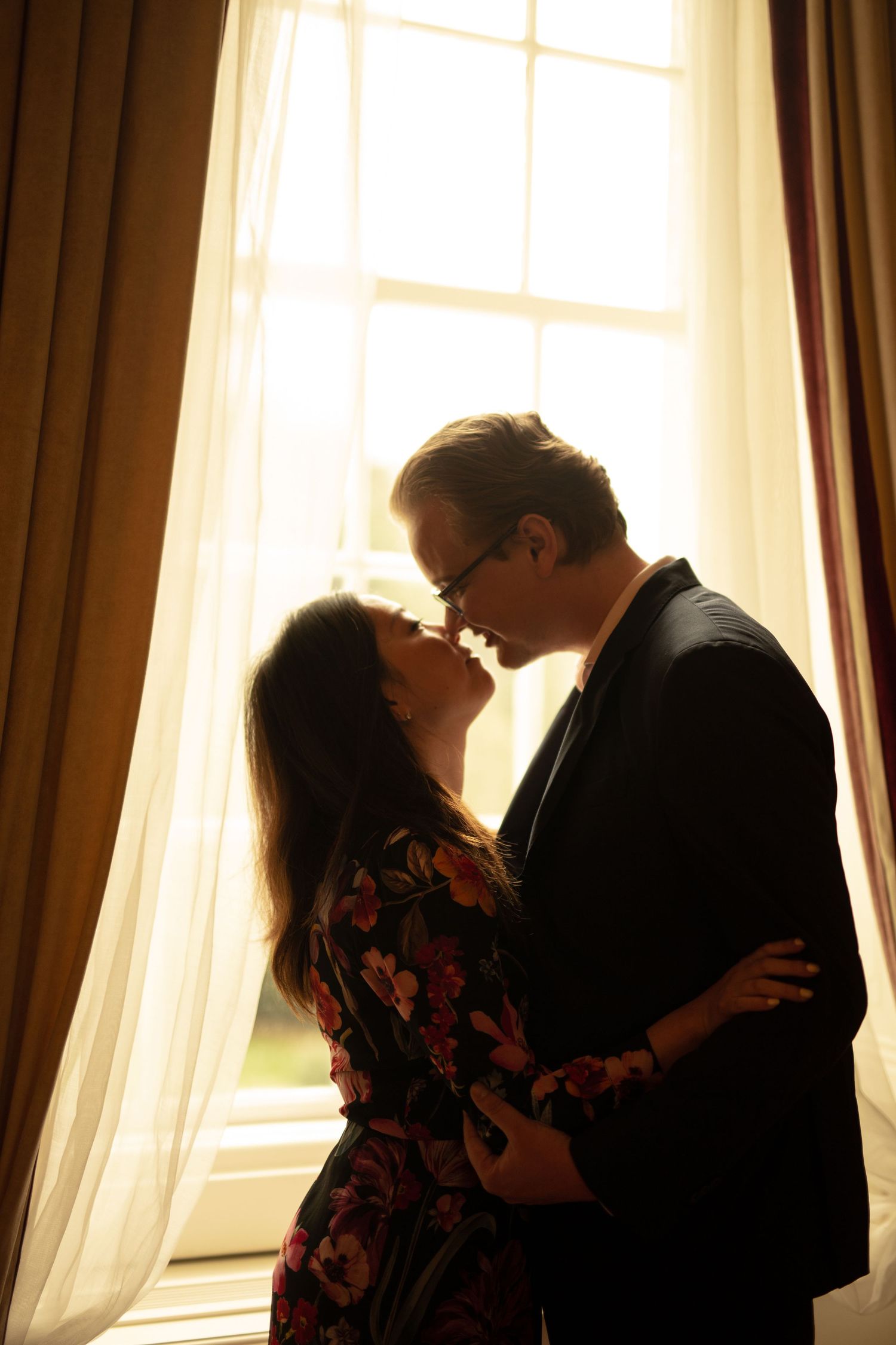 Romantic couple in silhouette sharing an intimate moment by a sunlit window with sheer curtains.
