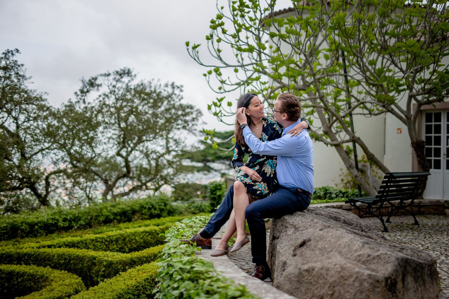 A couple shares tender engagement photos in a picturesque garden setting with stone walls and lush greenery.