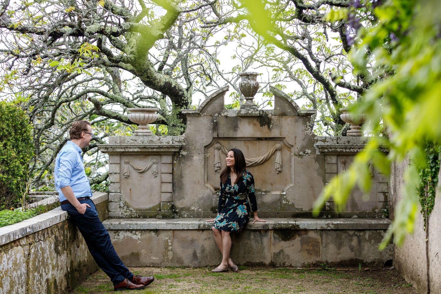 Several people sit and relax on an old stone wall surrounded by blooming spring trees and foliage in a garden setting.