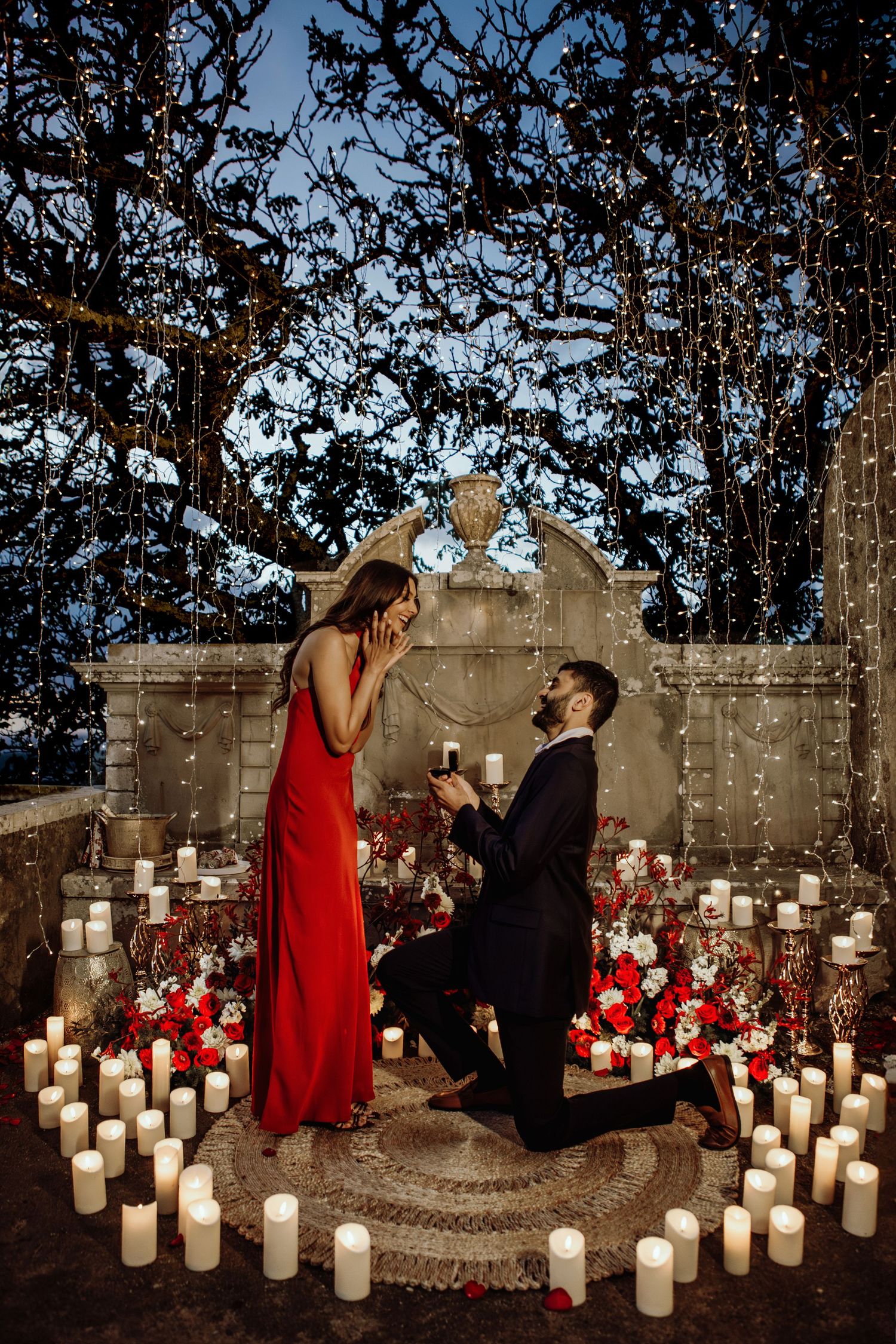 A romantic proposal scene in the evening surrounded by glowing white pillar candles against a stone wall.