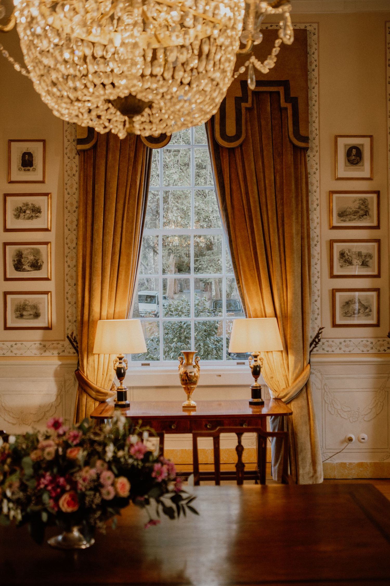 Elegant Victorian room interior with crystal chandeliers, gold curtains, and floral arrangements near windows.