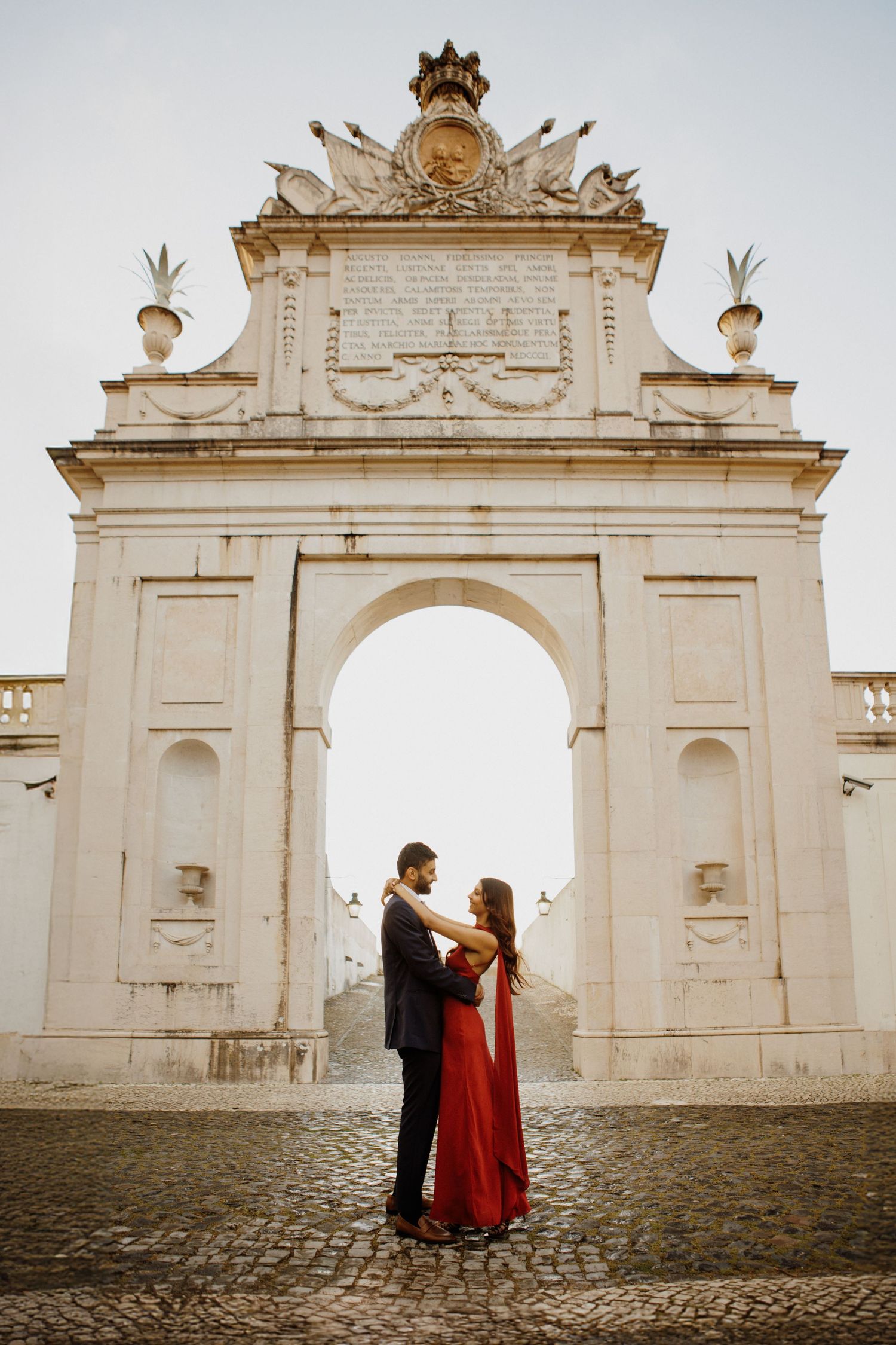 A couple stands embracing in front of an ornate white stone archway with decorative crown and architectural details.