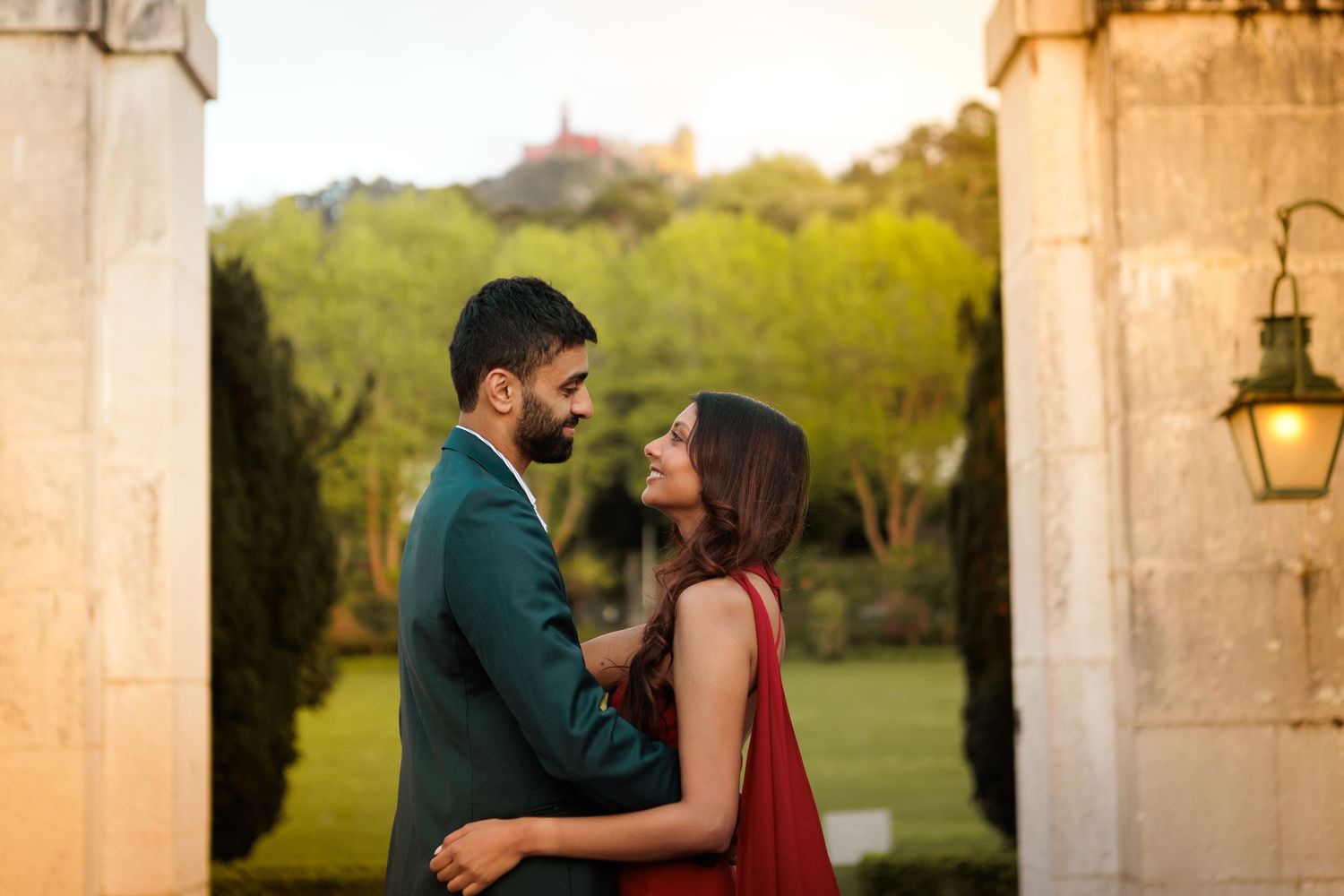 A couple in formal attire shares a romantic moment between stone pillars in an outdoor garden setting at sunset.