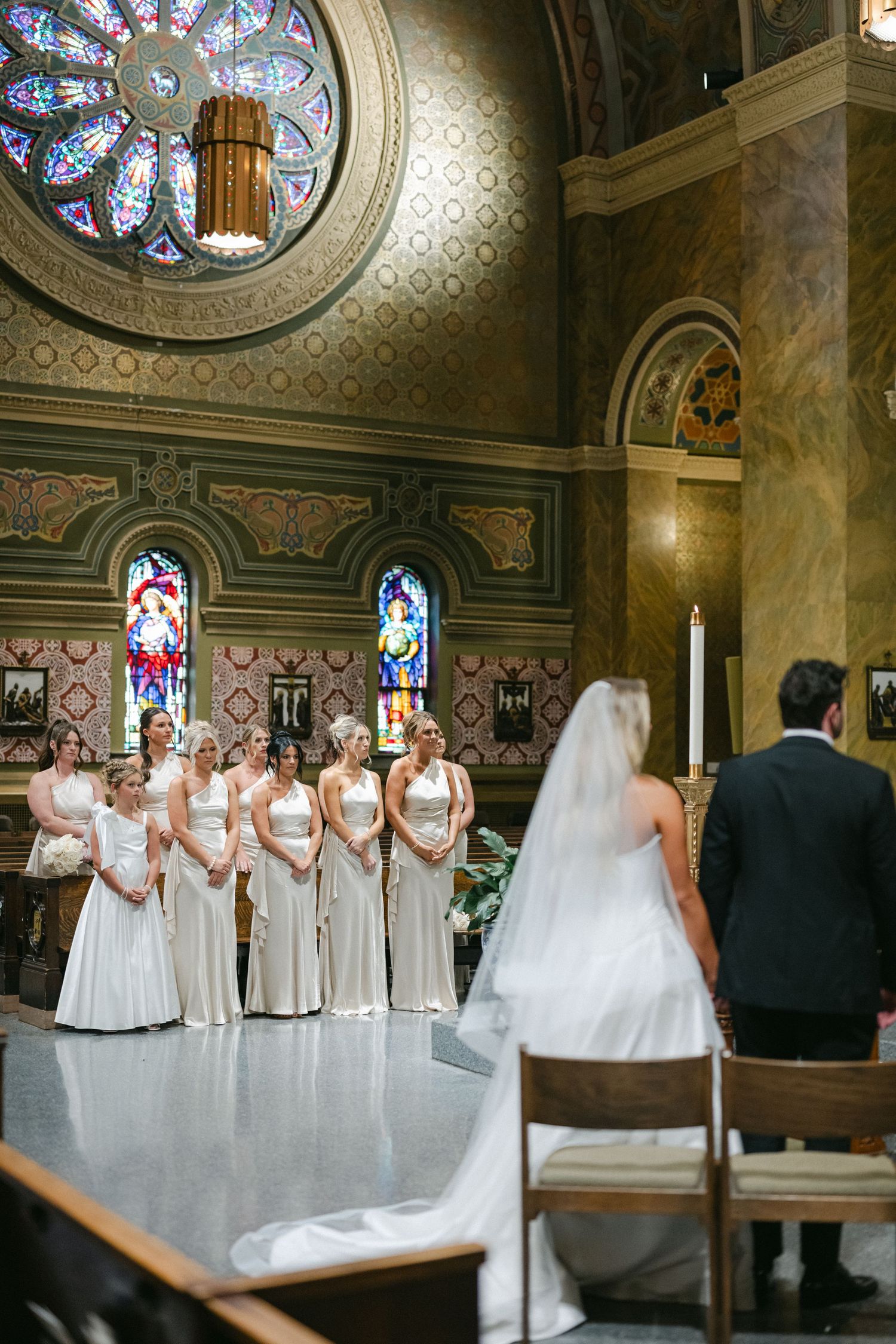 Wedding party stands in an elegant church interior with stained glass windows and ornate architectural details.