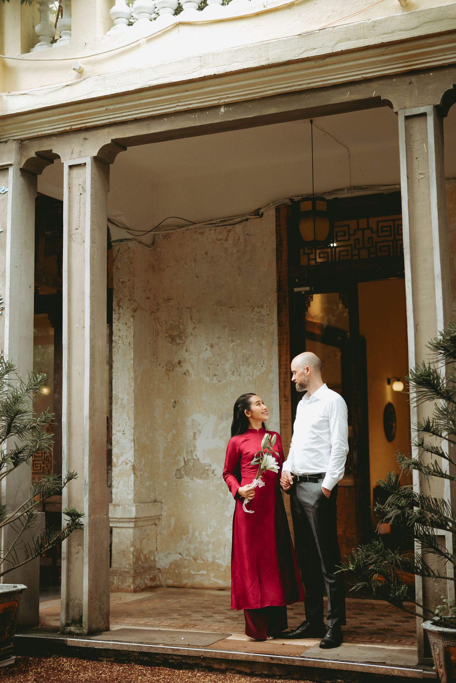 Couple in red ao dai and white shirt stand together in a vintage courtyard.
