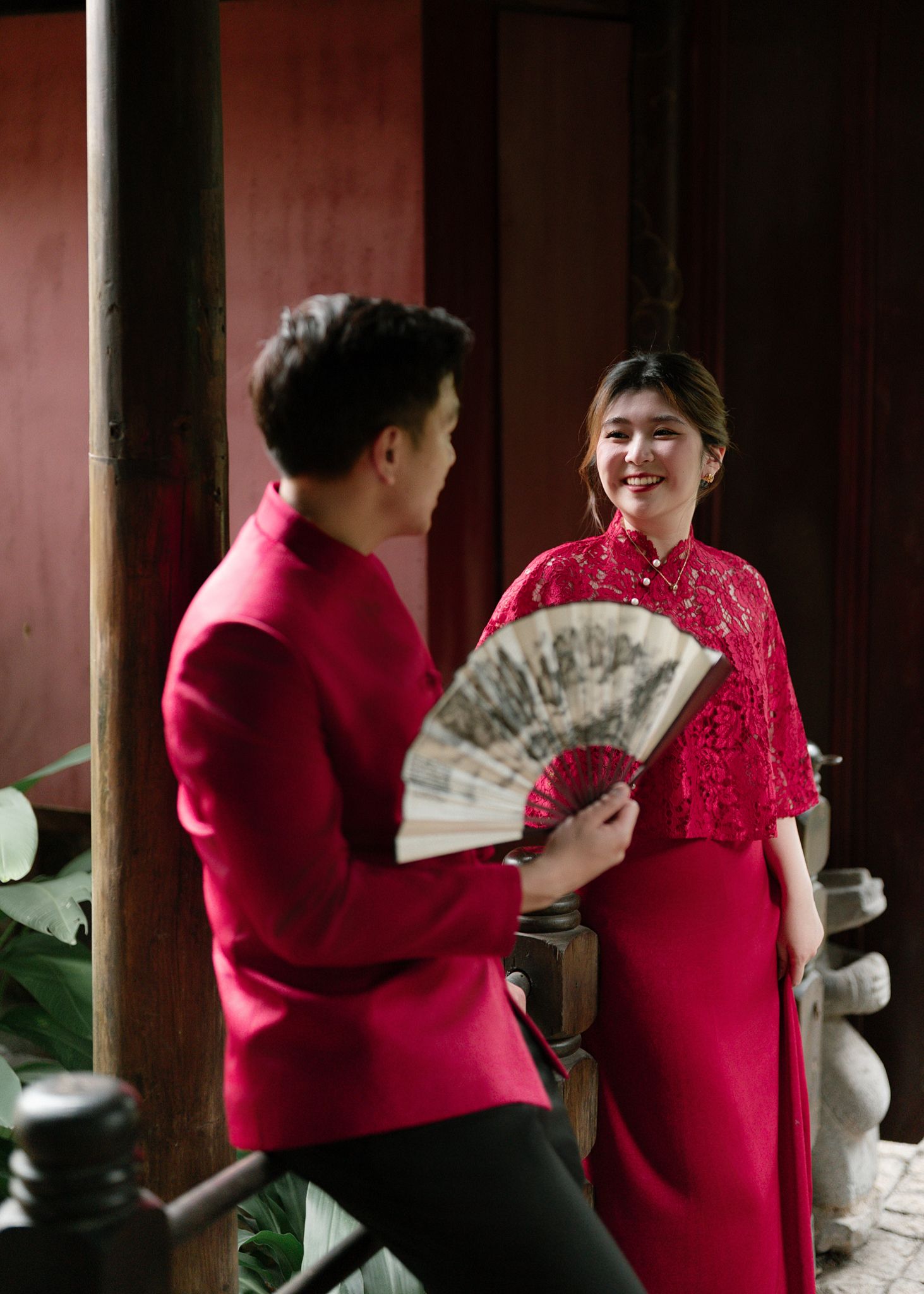 Couple in matching red traditional Vietnamese outfits share a joyful moment indoors.