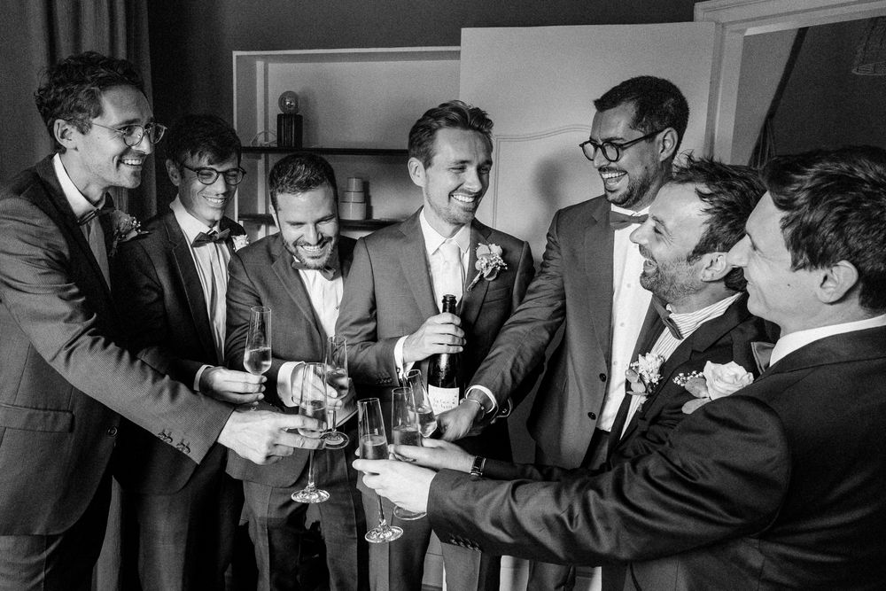 Moment chic et convivial a la fin des preparatifs du marie : coupe de champagne, sourires et complicite entre le marie et ses temoins.