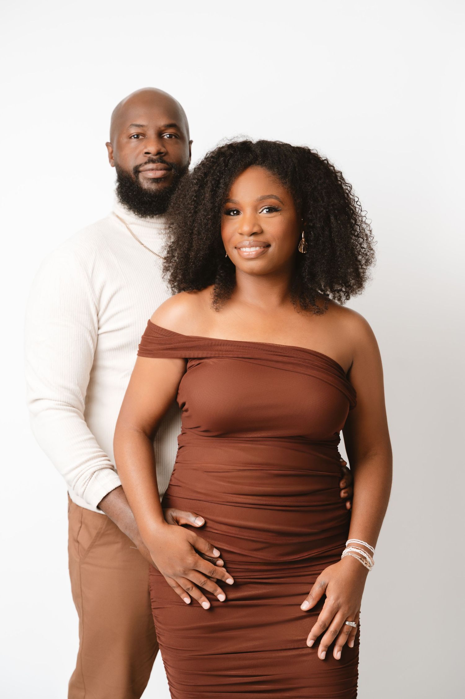 A couple poses together in matching brown and white outfits against a light background for a maternity photoshoot.