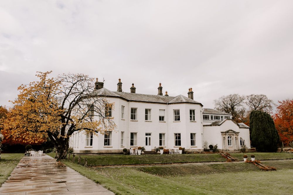A panoramic view of a classic white colonial mansion with symmetrical windows and autumn trees in the background.