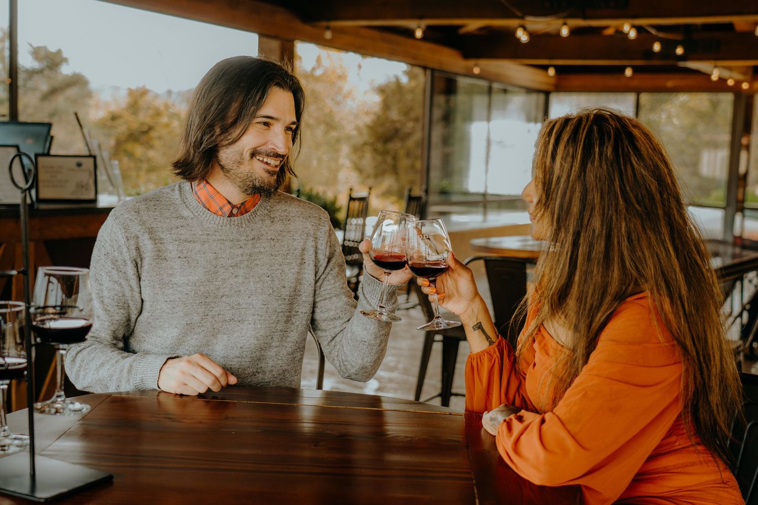 Couple enjoys wine tasting together in rustic venue with string lights overhead.