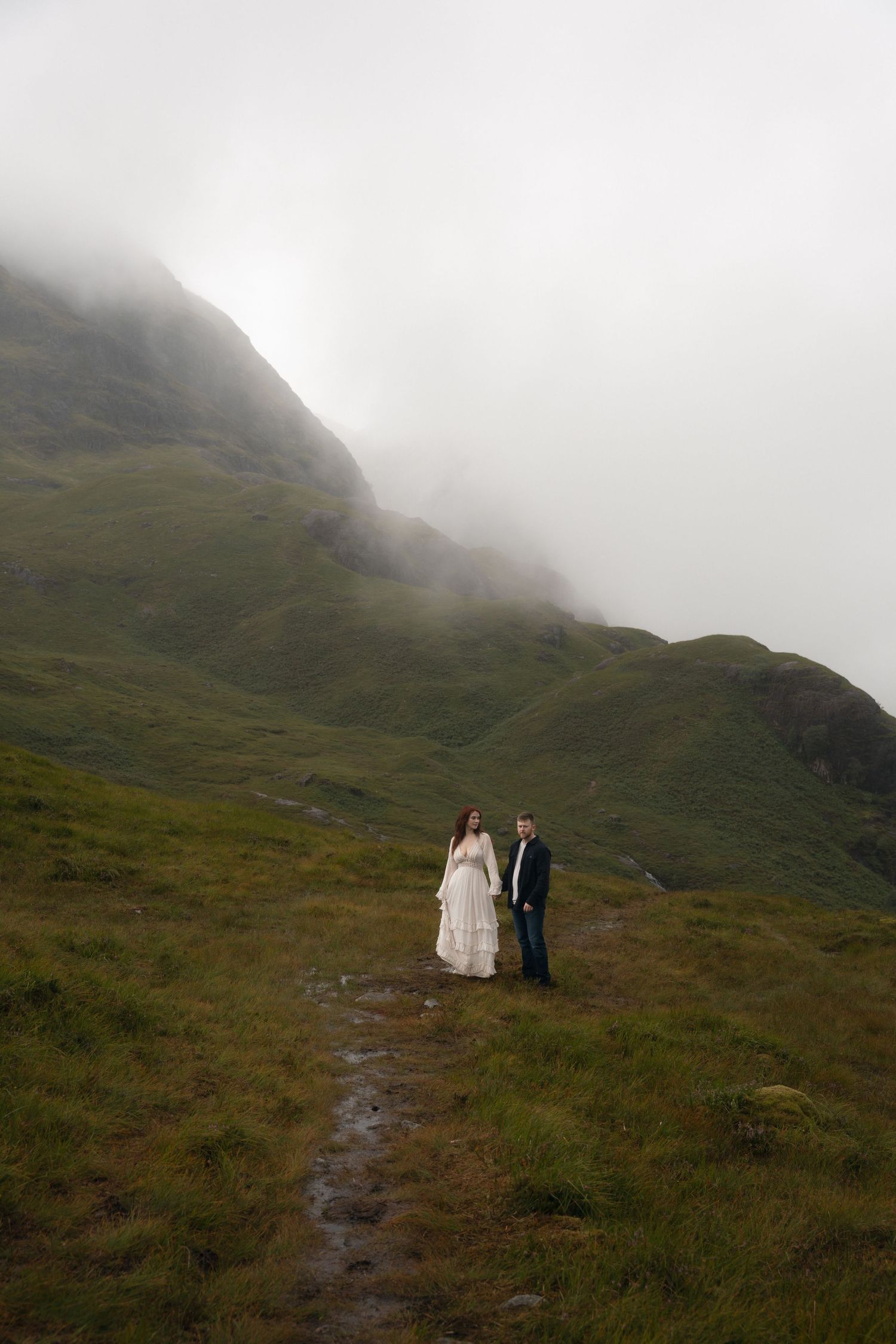 Two figures walk along a misty mountain trail surrounded by green slopes and foggy peaks in Scotland.
