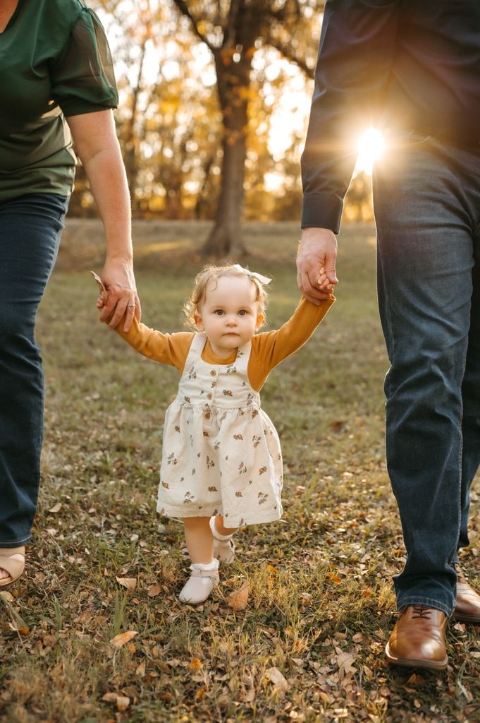 A toddler in a floral dress holds hands with adults while walking through fallen leaves at sunset.