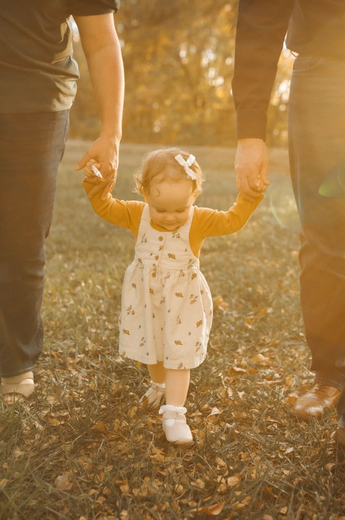 Small feet in white socks walking through autumn leaves while holding hands with an adult in golden sunlight.