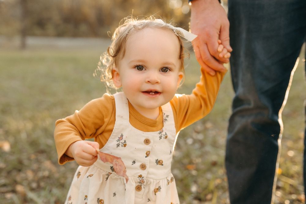 A toddler in a white patterned dress and yellow cardigan smiles while being supported by adult hands.