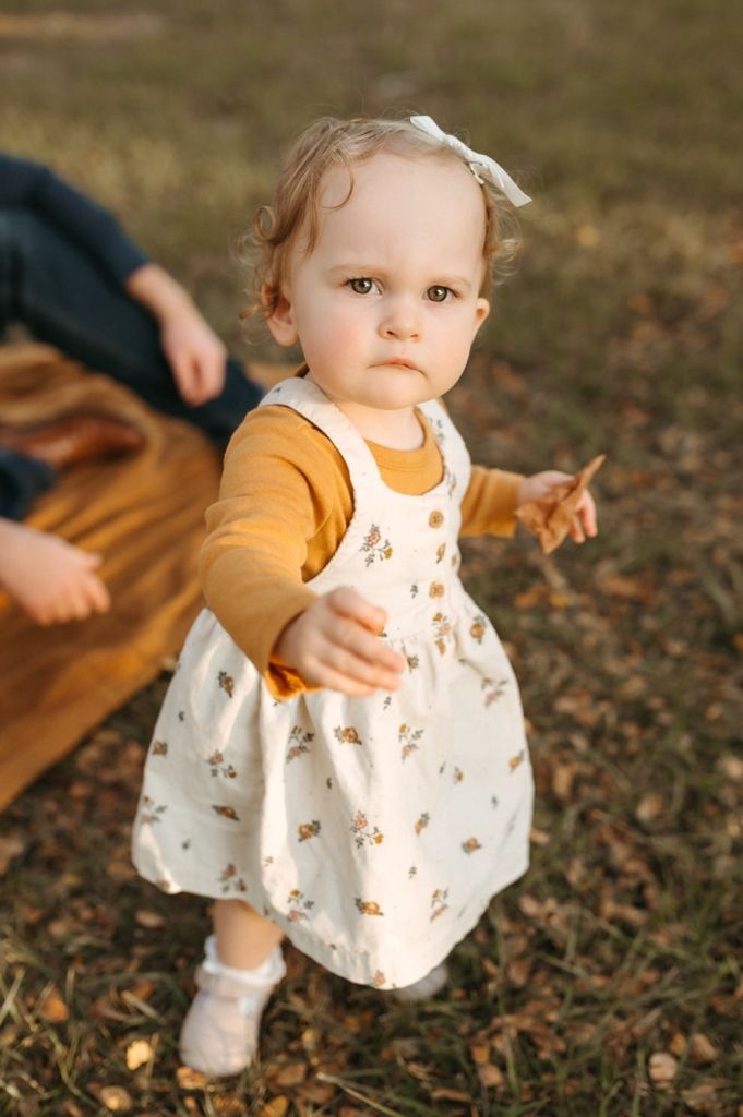 A small child in a white dress and yellow cardigan explores fallen leaves during golden hour.