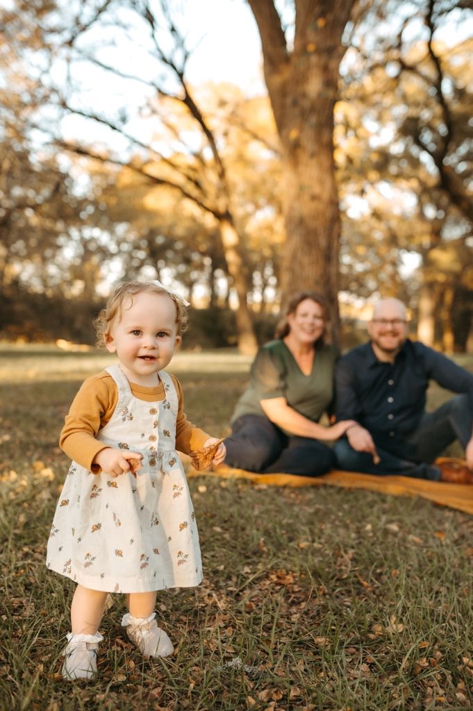 A toddler in a floral dress and mustard cardigan plays on autumn grass while family relaxes in background.