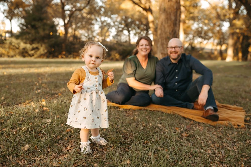 A toddler in a white patterned dress and yellow sweater explores a leaf-covered lawn at sunset.
