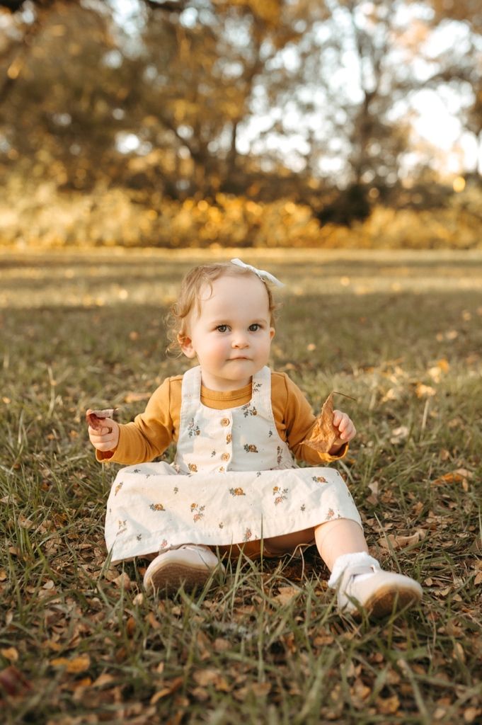 A small child sits on autumn grass wearing a floral dress and brown cardigan in warm golden light.