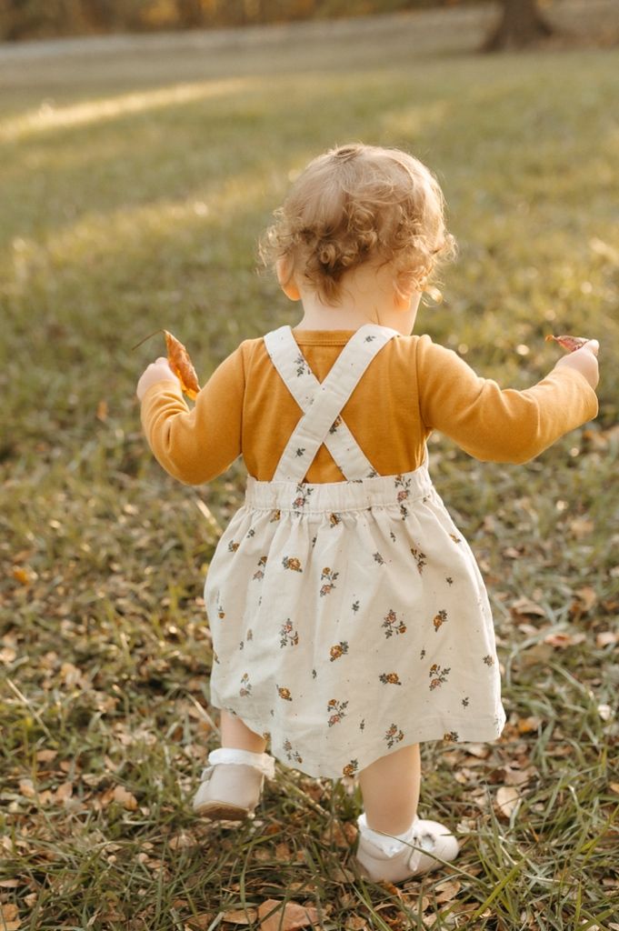 Back view of a baby wearing a white dress with crossed straps and yellow cardigan walking on grass.