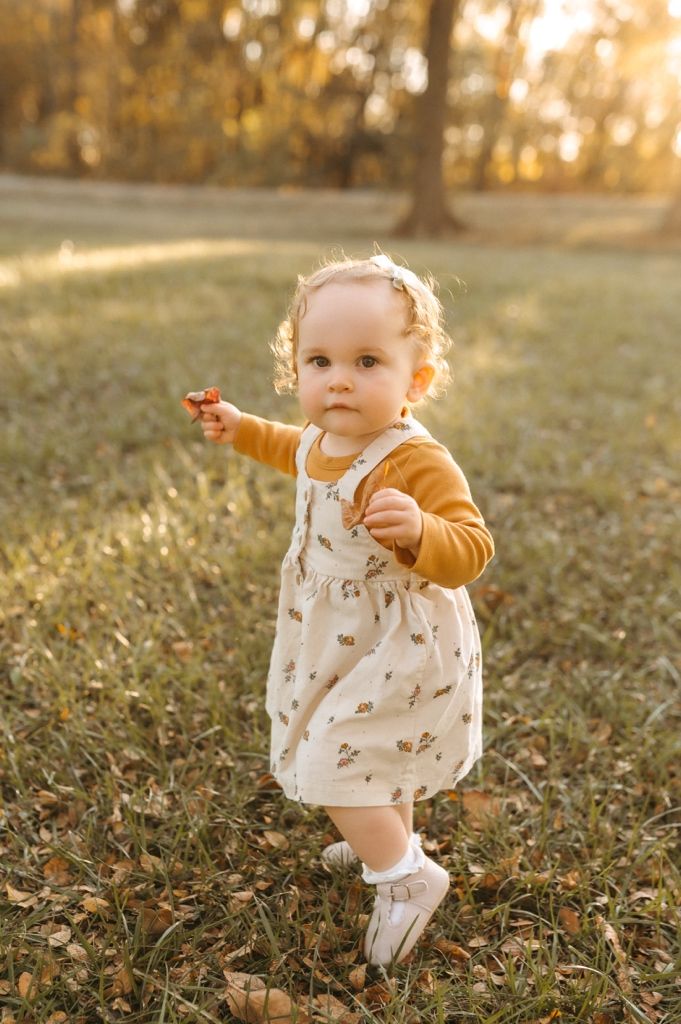 A toddler in a floral dress and mustard cardigan points while standing in a sunlit autumn park.