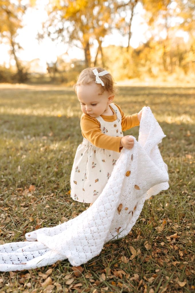 A toddler in a mustard sweater and patterned dress holds a delicate white lace blanket in an autumn setting.