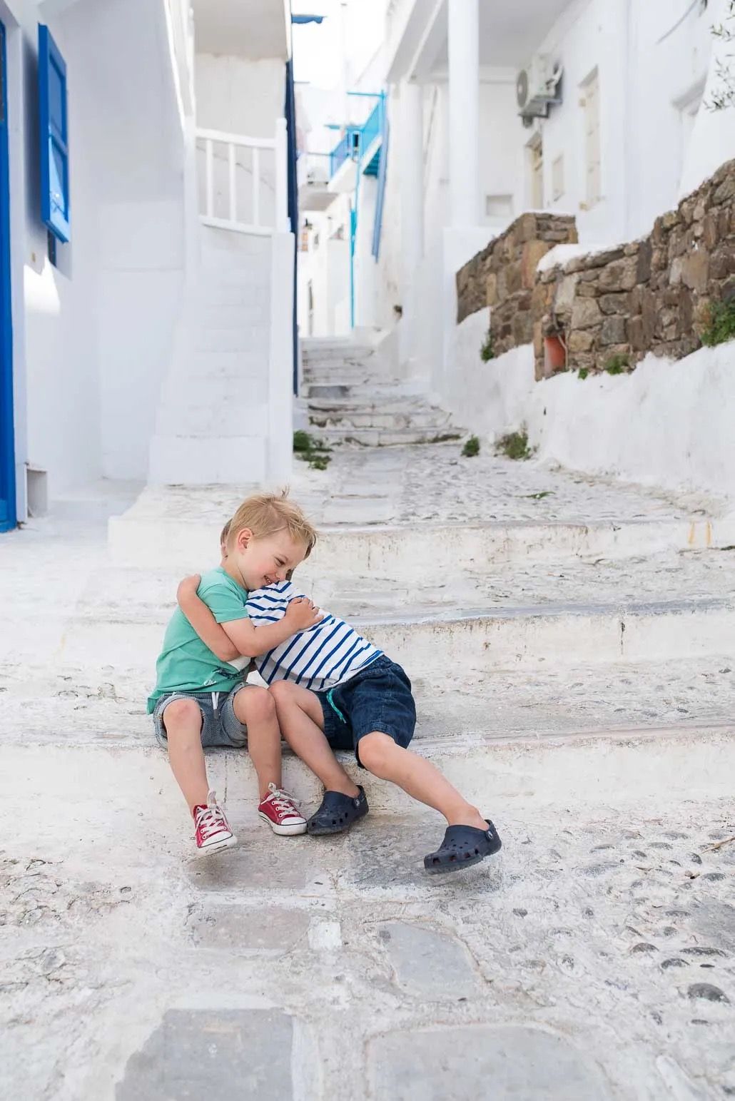Two young children sitting on white stone steps in a bright Greek island alleyway.