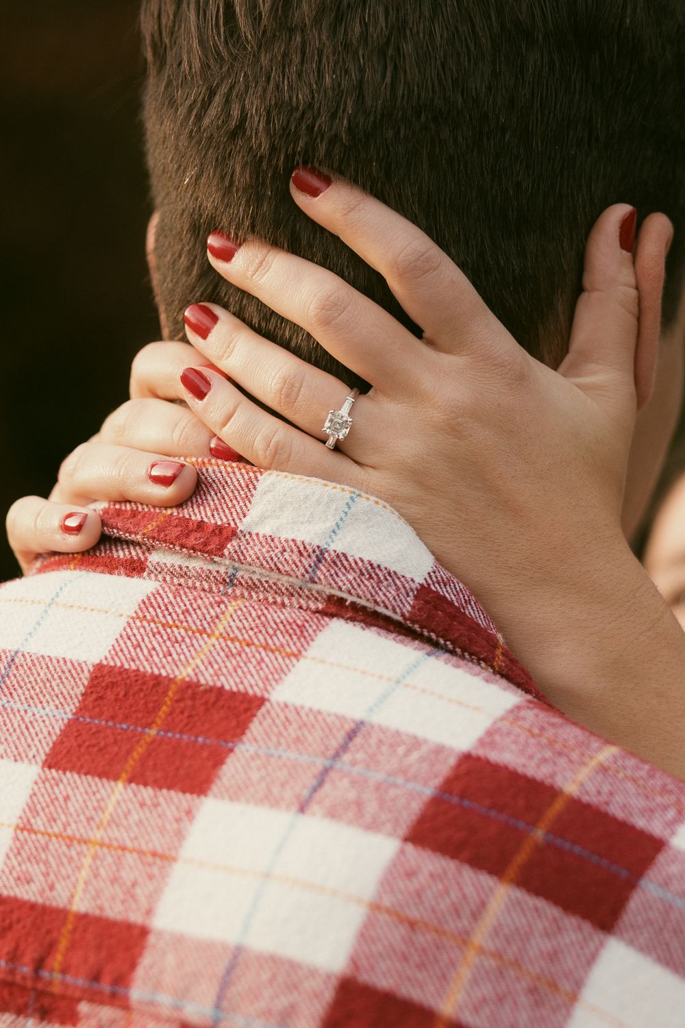 Close-up of hands with engagement ring resting on red and white plaid fabric.