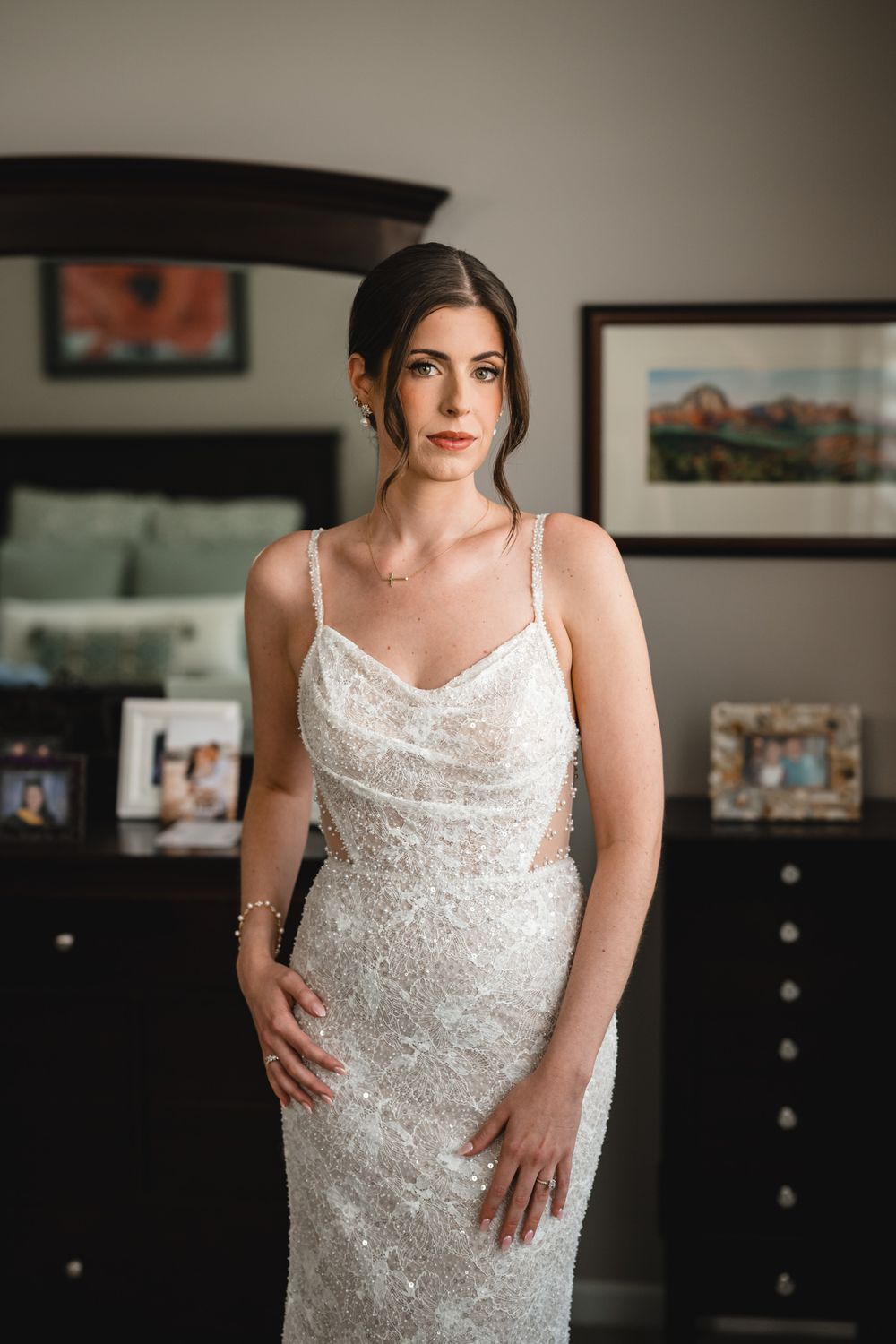A bride poses in a stunning sequined white wedding gown with spaghetti straps in a bedroom setting.