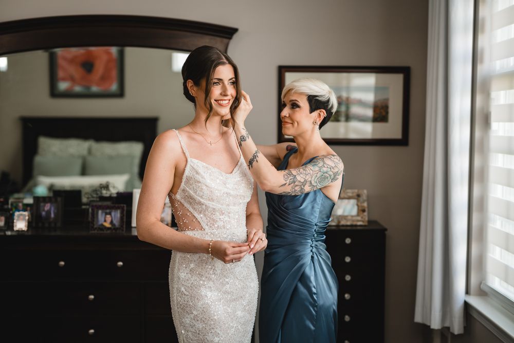 A bride in a sparkling white gown shares an intimate moment with someone in a navy dress near a window.