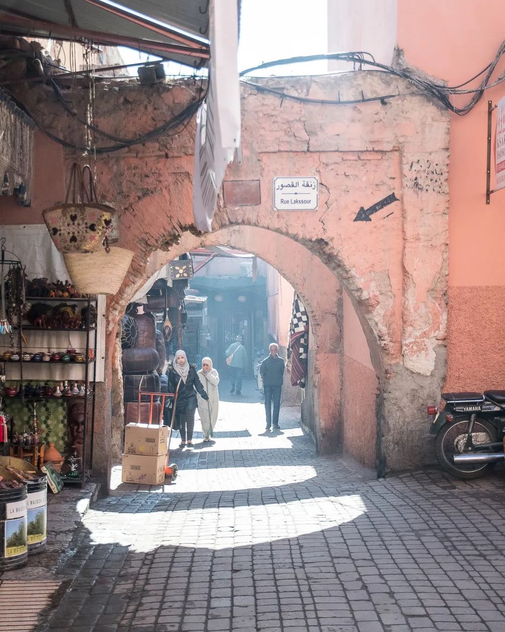 A sunlit Moroccan market street with shops lining an archway and people walking through.