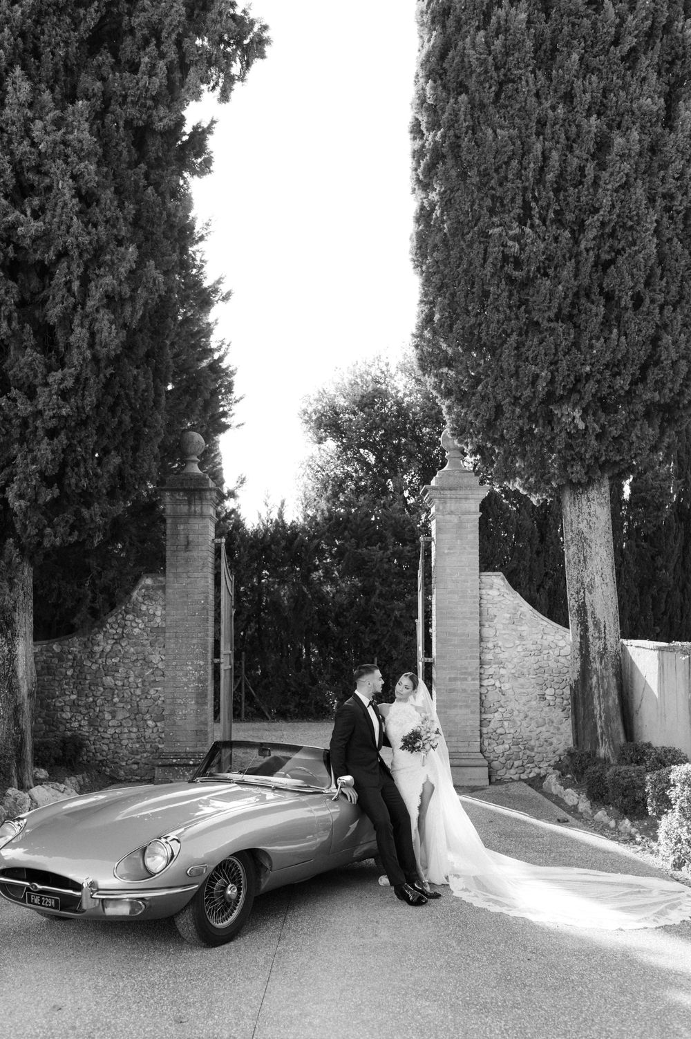Black and white photo of newlyweds posing with vintage Jaguar E-Type car by stone pillars and cypress trees.