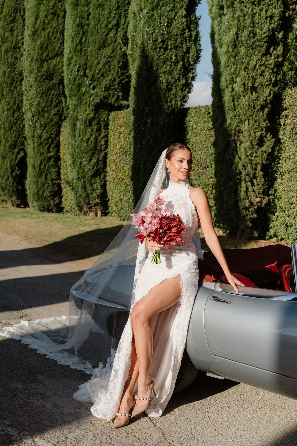 A bride in a white dress poses elegantly with a red bouquet next to a silver car on a cypress tree-lined path.