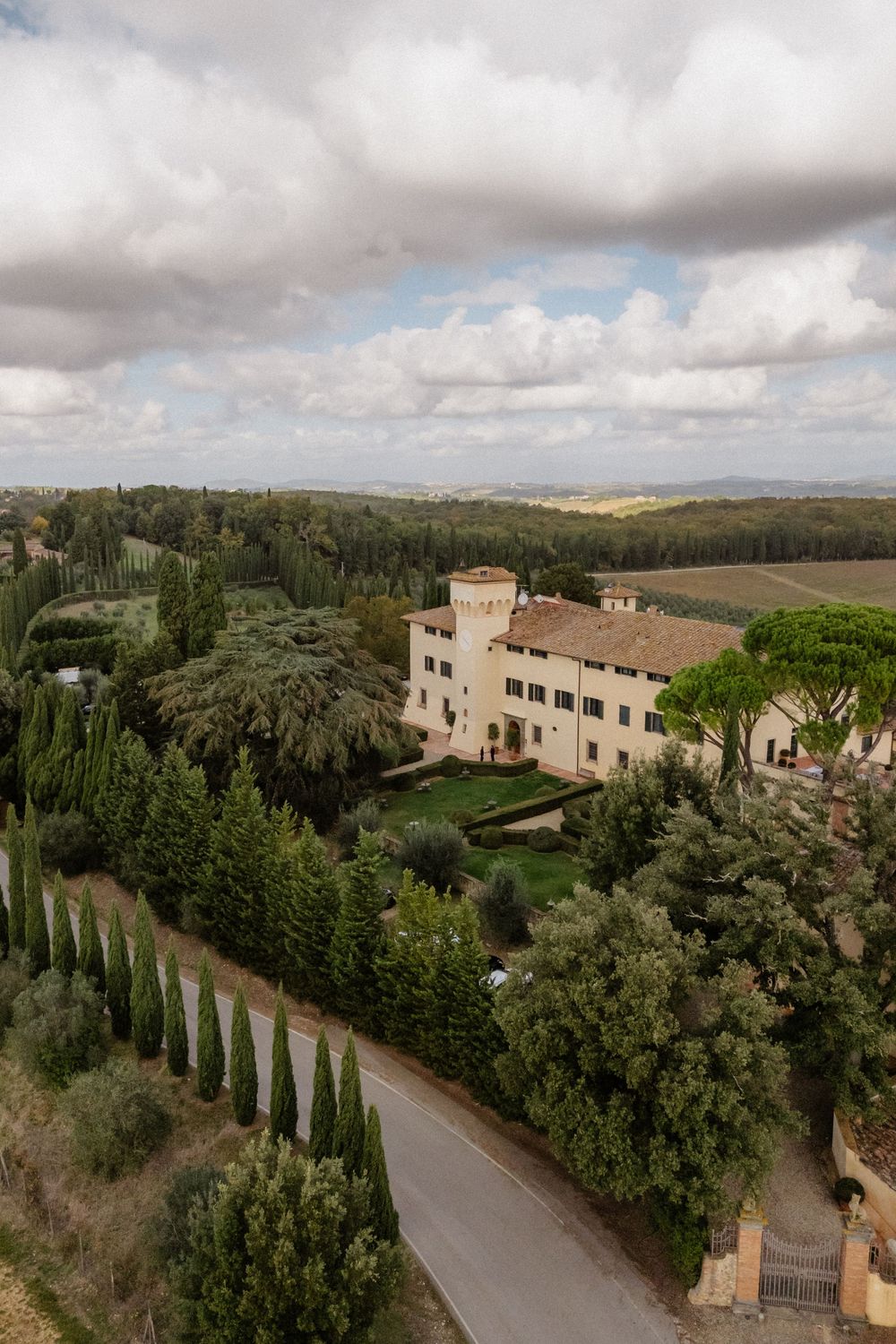Aerial panoramic view of a Tuscan villa surrounded by cypress trees and lush gardens under cloudy skies.