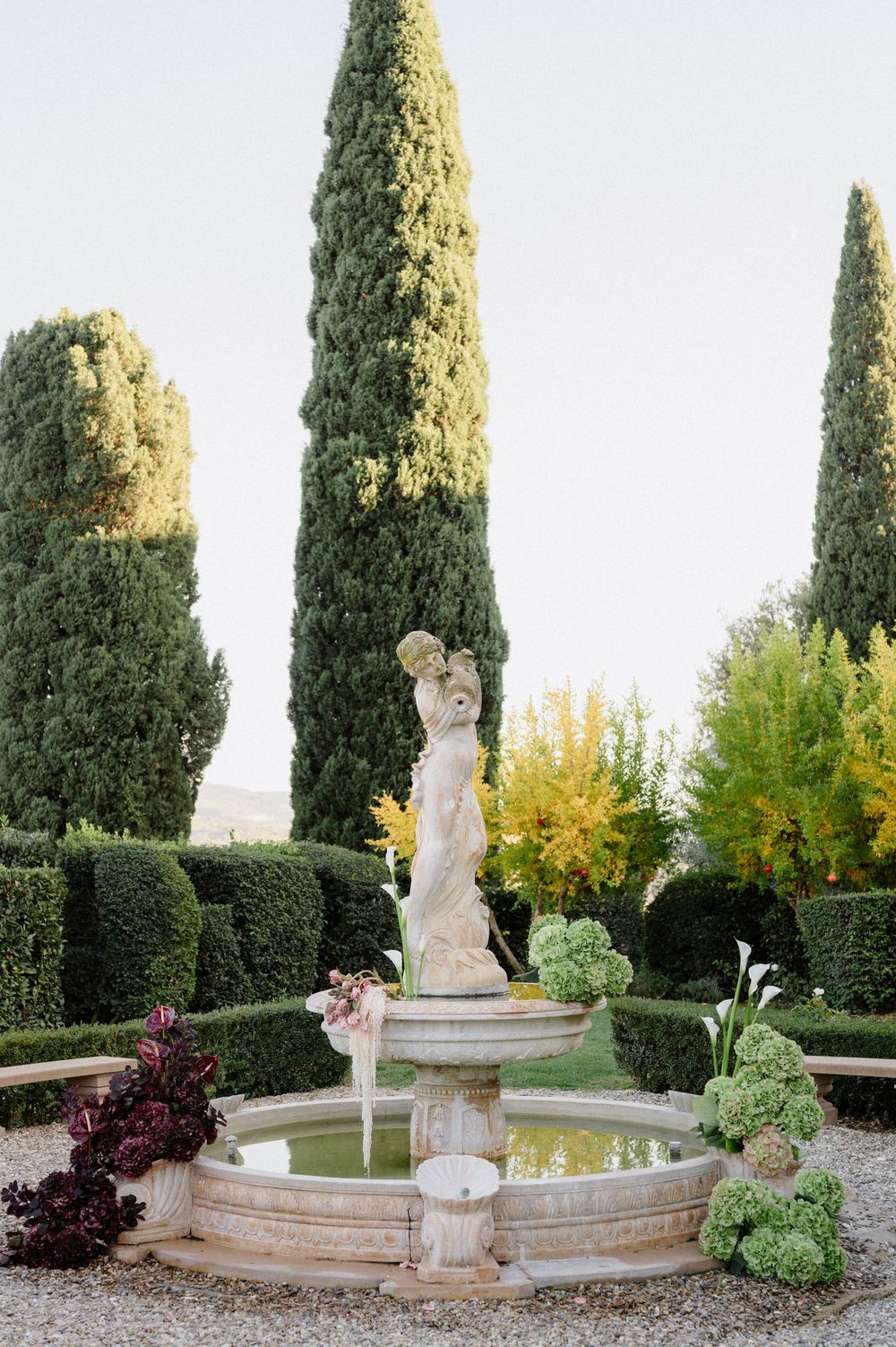 Elegant garden fountain with cherub statue surrounded by tall cypress trees and manicured hedges in a formal setting.