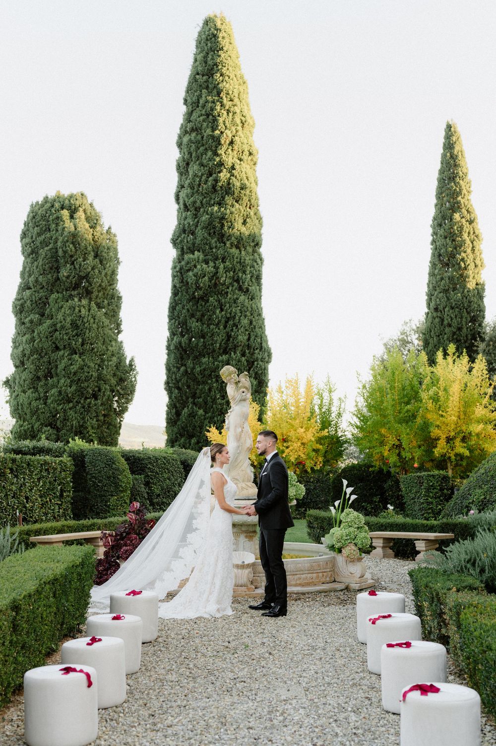 A wedding ceremony takes place on a gravel path lined with white lanterns and tall cypress trees in an elegant garden setting.