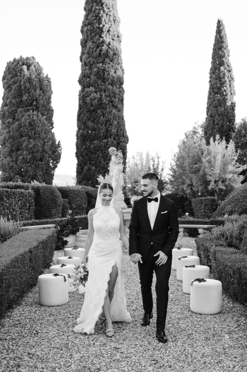 Black and white wedding photo series showing a couple posing in a formal garden with cypress trees and white decor elements.