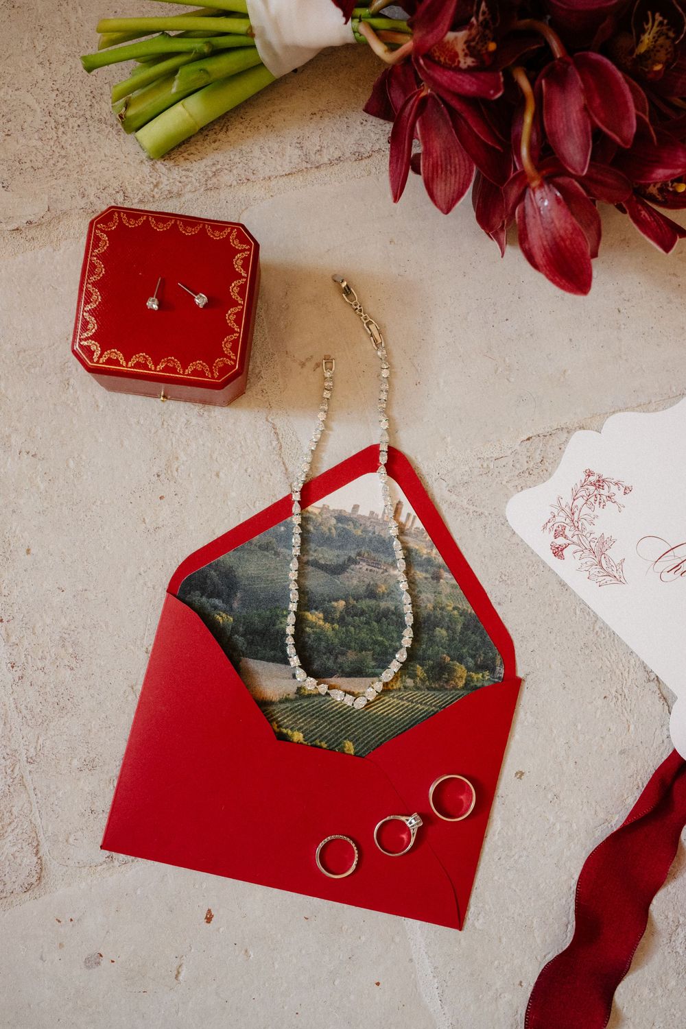 Wedding rings and pearl necklace displayed on a red envelope with deep red flowers against a textured beige surface.