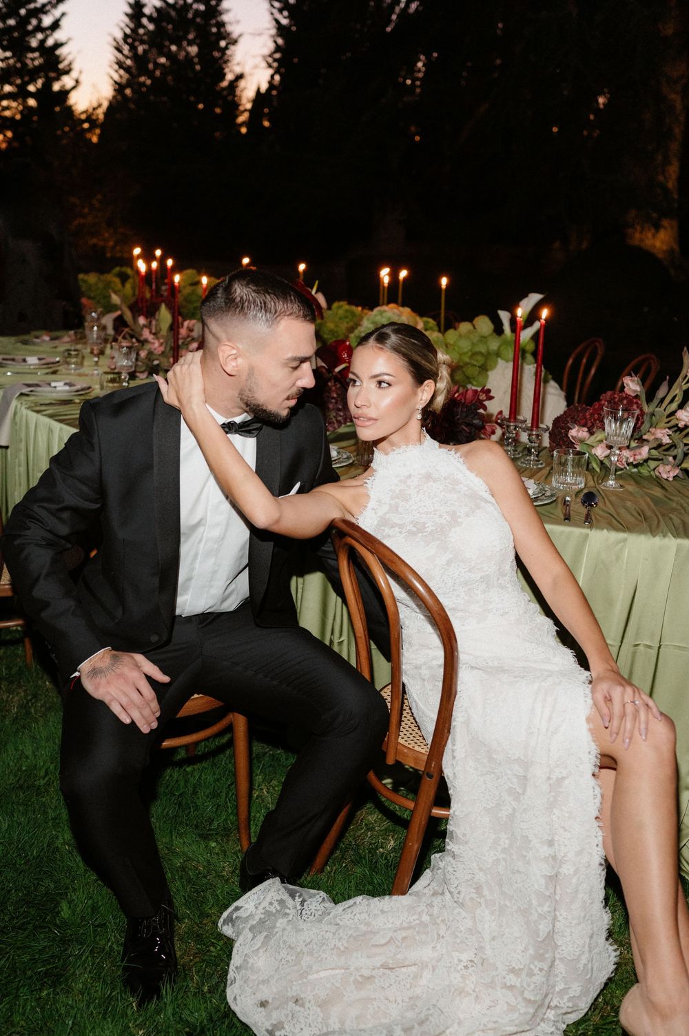 Romantic wedding couple poses together on vintage chair during outdoor evening reception with elegant table settings.