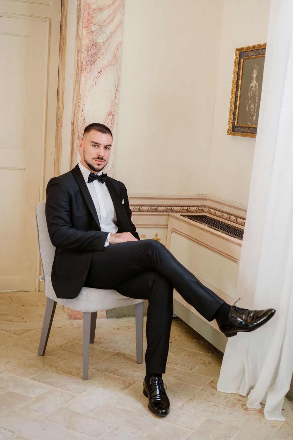 A groom in a black tuxedo sits casually on a gray chair in an elegant room with light walls.