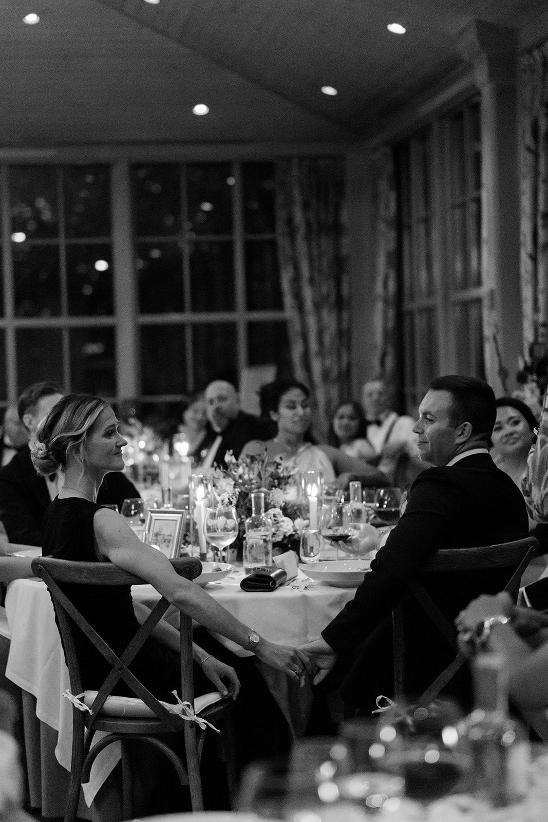 Wedding guests seated at elegant dining tables in a dimly lit reception hall with large windows and ambient lighting.