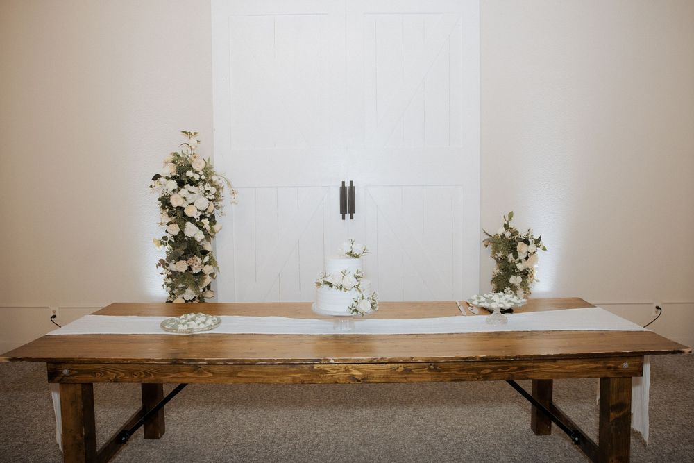 A rustic wooden dining table with white floral arrangements and black metal legs sits against a white wall.
