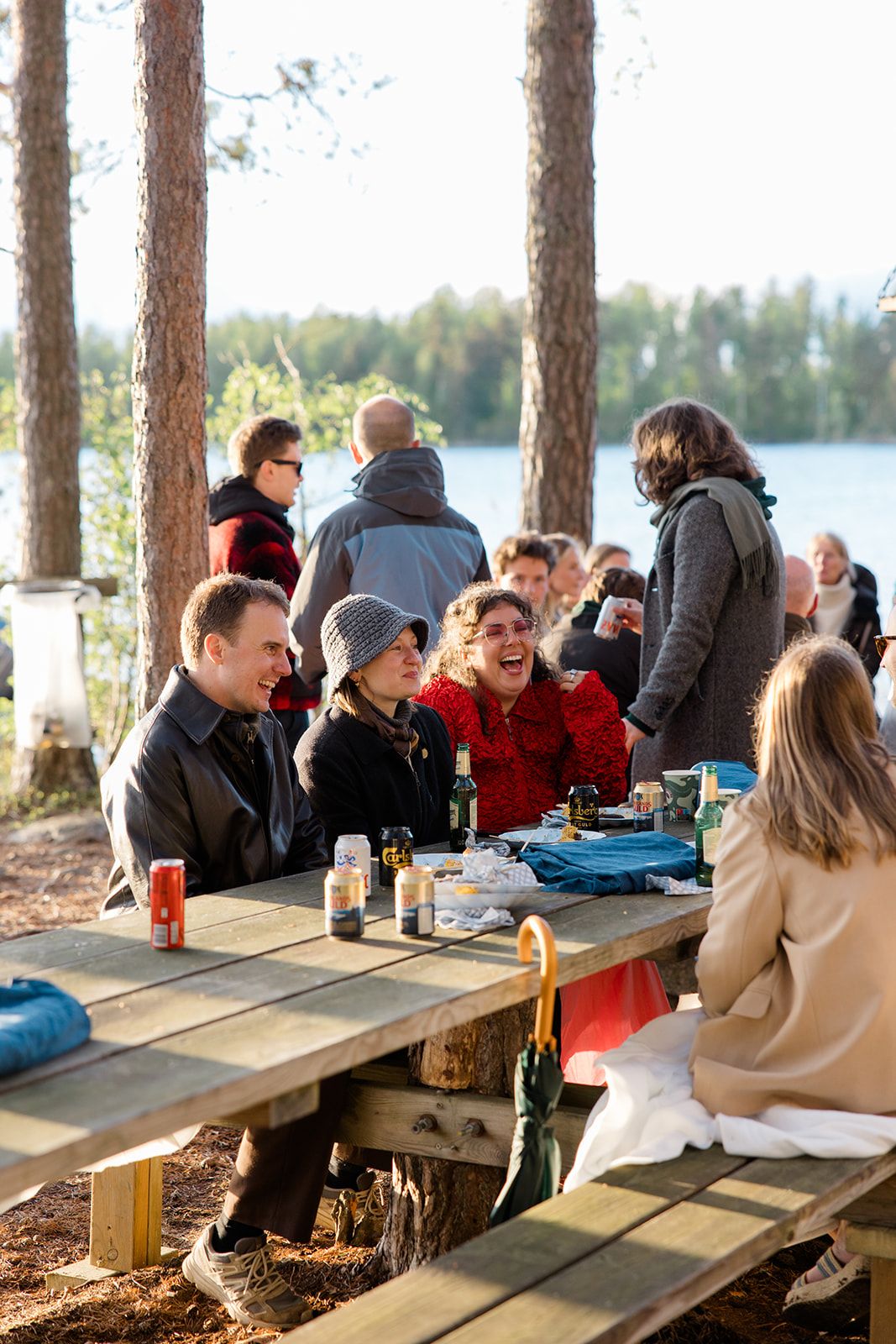 Group of people sitting at long wooden picnic tables in a forest setting beside a lake on a sunny day.