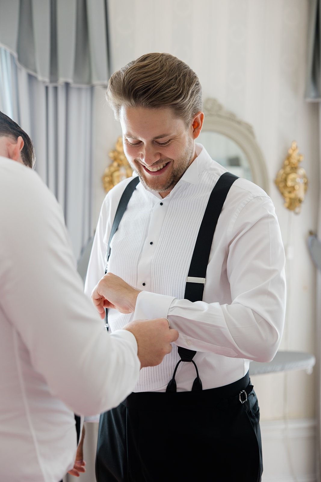 A person in a white dress shirt and black suspenders smiles while getting dressed for a formal occasion.