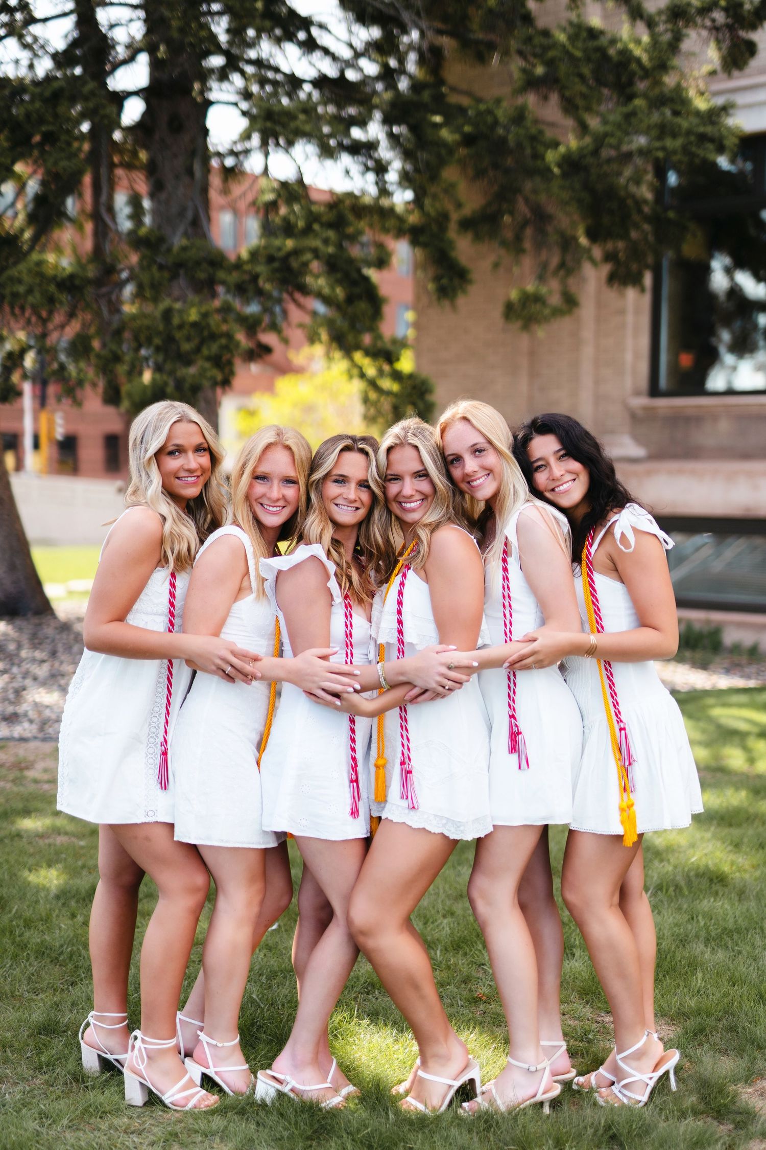 A group in white dresses with colorful sashes poses together on grass near a building in three similar shots.