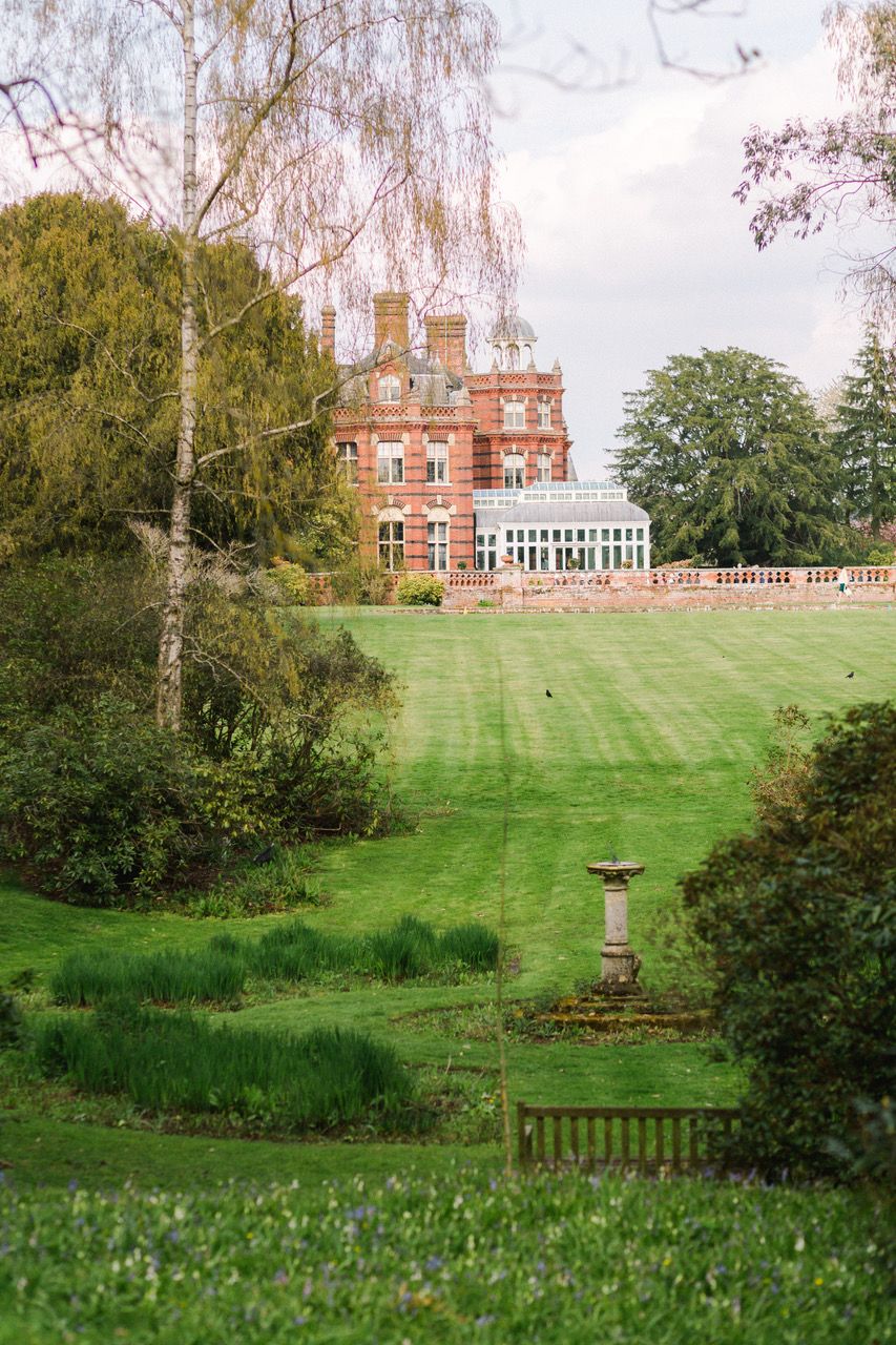 A panoramic view of a grand red brick mansion set atop a sloping green lawn with birch trees and garden features at The Elvetham