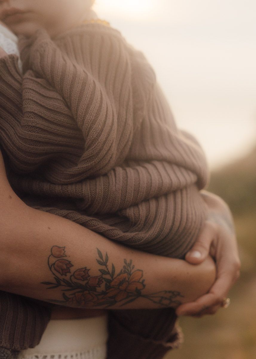 Person in brown knit sweater showing floral arm tattoo against soft sunlit background.