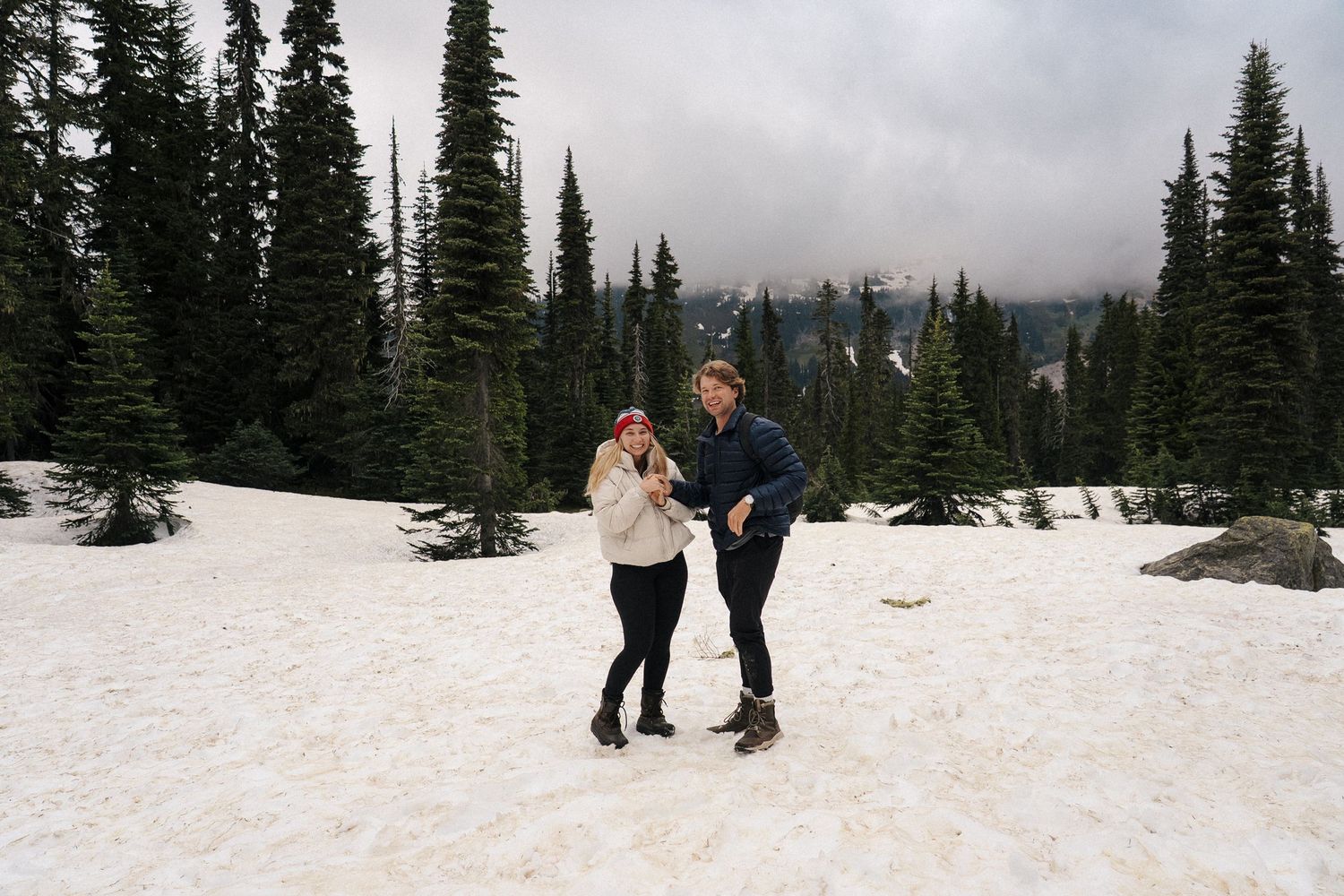 A couple walks together through a snowy mountain landscape surrounded by tall evergreen trees during winter.